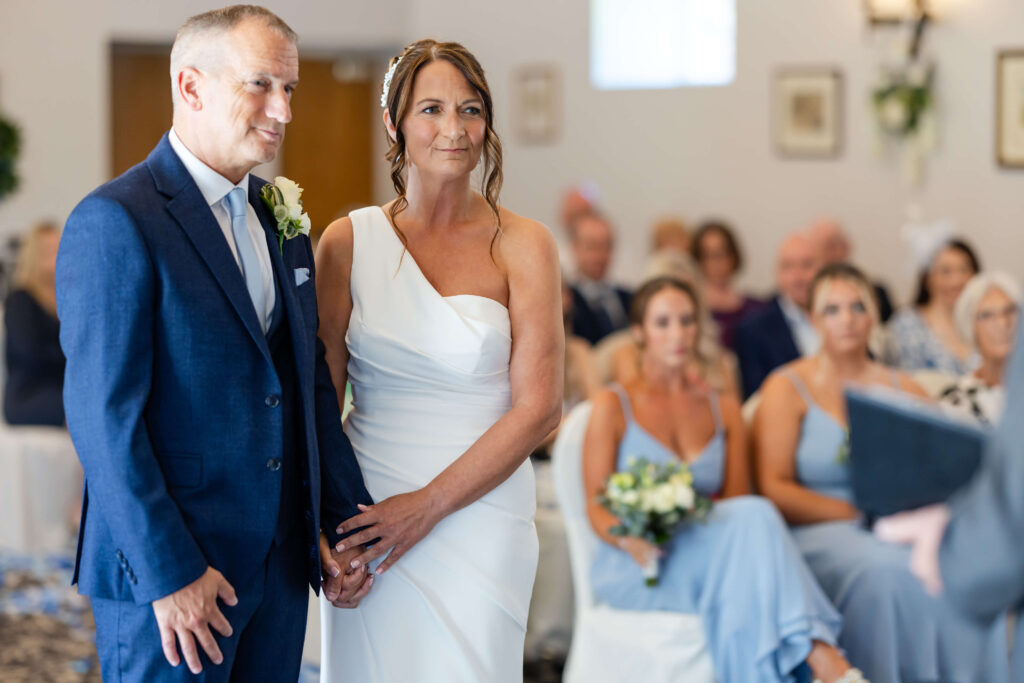 A bride in a white dress and a groom in a blue suit stand holding hands at an indoor wedding ceremony, with seated guests in the background. Copyright Somerside Photography Ltd // www.somersidephotography.com