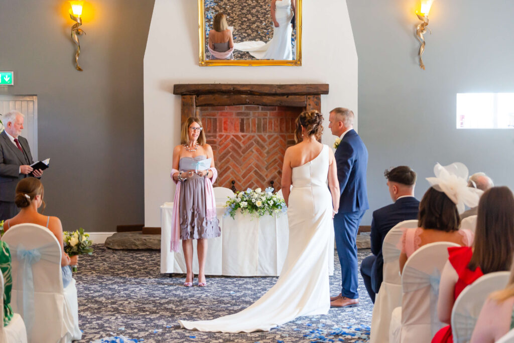 A bride and groom stand at the altar during a wedding ceremony, facing the officiant, with guests seated and watching in a decorated indoor venue. Copyright Somerside Photography Ltd // www.somersidephotography.com