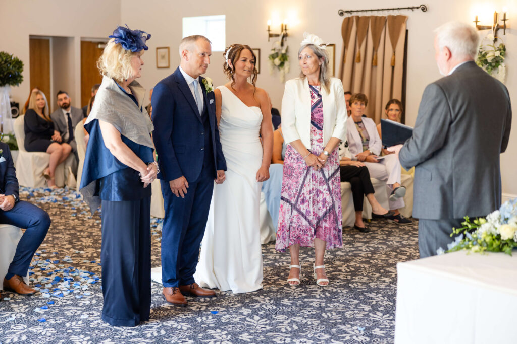A bride and groom stand together at an indoor wedding ceremony, accompanied by two women, likely their mothers, as a man officiates. Guests are seated and watching in the background. Copyright Somerside Photography Ltd // www.somersidephotography.com