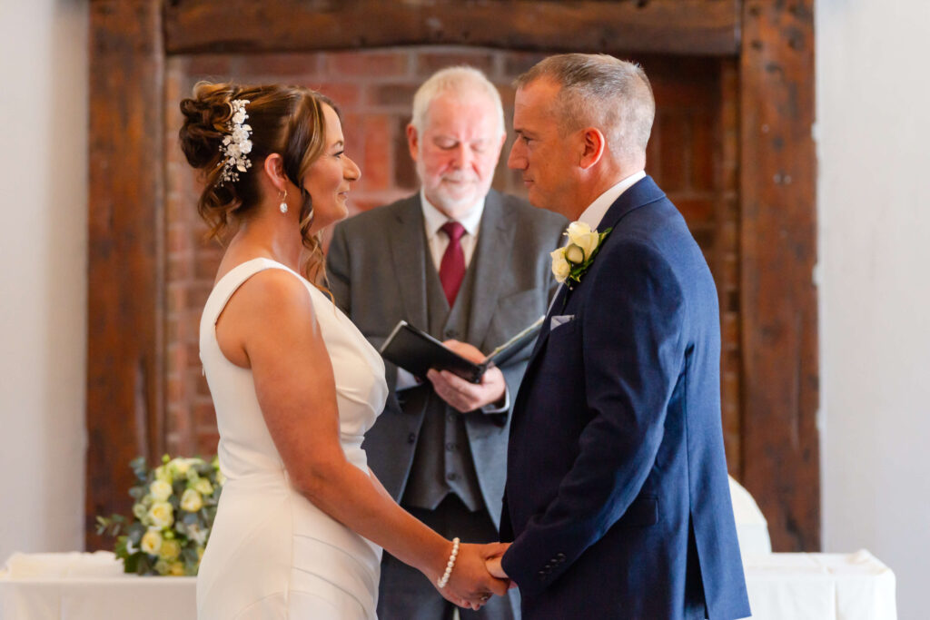 A bride and groom stand facing each other and holding hands during a wedding ceremony, with an officiant standing behind them. Copyright Somerside Photography Ltd // www.somersidephotography.com