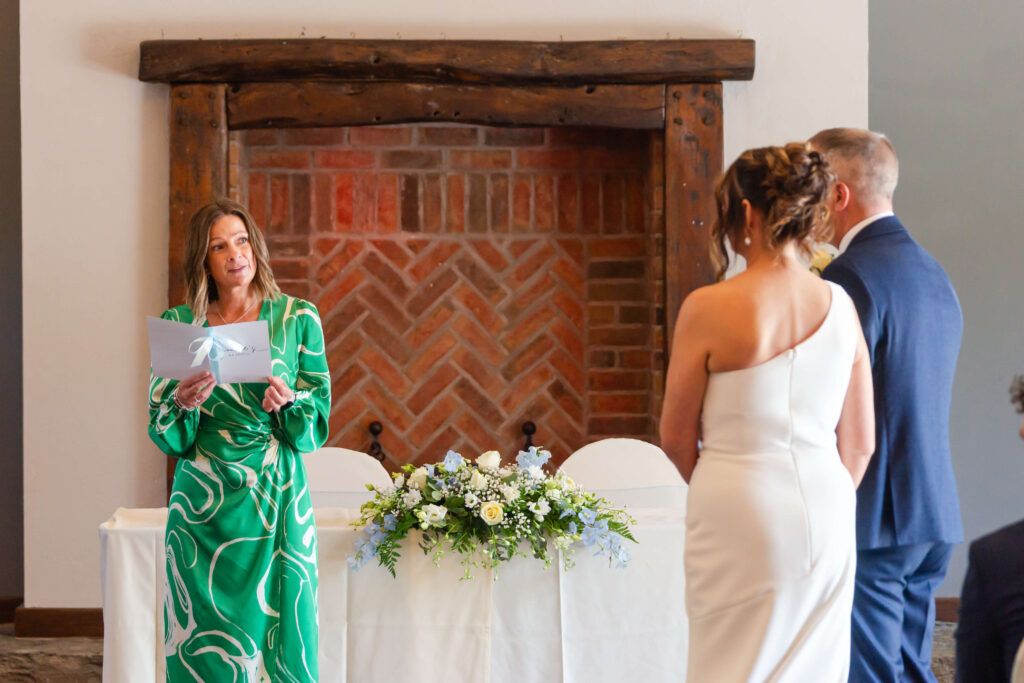 A woman in a green dress reads from a folder, facing a bride and groom standing in front of a table with floral arrangements and a brick fireplace. Copyright Somerside Photography Ltd // www.somersidephotography.com