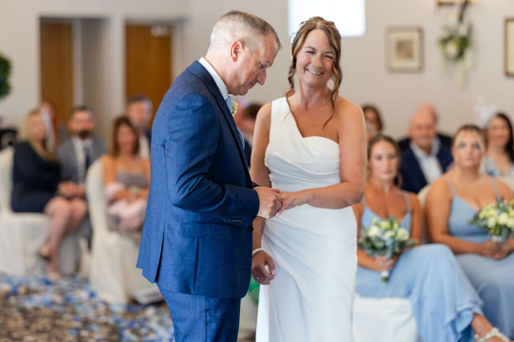 A bride and groom exchange rings during a wedding ceremony, with seated guests watching in the background. Copyright Somerside Photography Ltd // www.somersidephotography.com