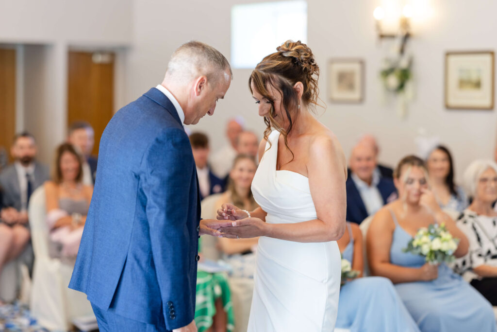 A bride and groom exchange rings during a wedding ceremony, with seated guests watching in the background. Copyright Somerside Photography Ltd // www.somersidephotography.com