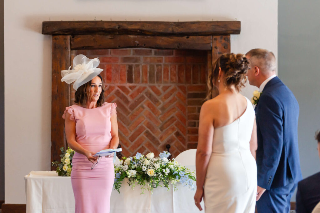 A woman in a pink dress and large white hat stands at a table with flowers, facing a couple dressed in formal attire, likely during a wedding ceremony. Copyright Somerside Photography Ltd // www.somersidephotography.com