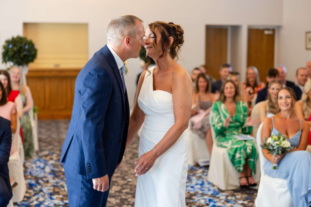 A bride and groom stand facing each other, about to kiss, during a wedding ceremony with seated guests watching in the background. Copyright Somerside Photography Ltd // www.somersidephotography.com