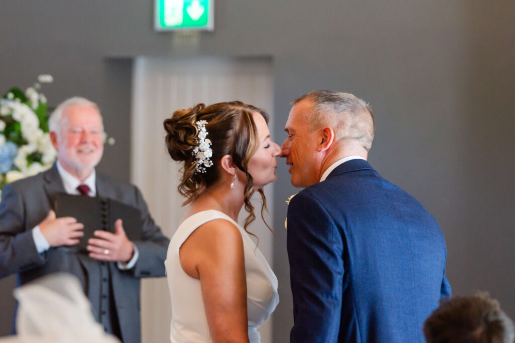 A bride and groom lean in for a kiss during their wedding ceremony, with an officiant smiling in the background. Copyright Somerside Photography Ltd // www.somersidephotography.com