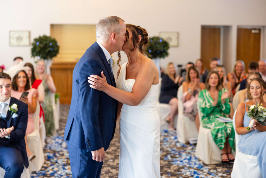 A bride and groom kiss at the altar during their wedding ceremony, with guests seated and applauding in the background. Copyright Somerside Photography Ltd // www.somersidephotography.com