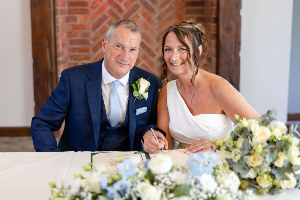 A man in a suit and a woman in a white dress sit together at a table, smiling, with a bunch of flowers and a document in front of them. Copyright Somerside Photography Ltd // www.somersidephotography.com