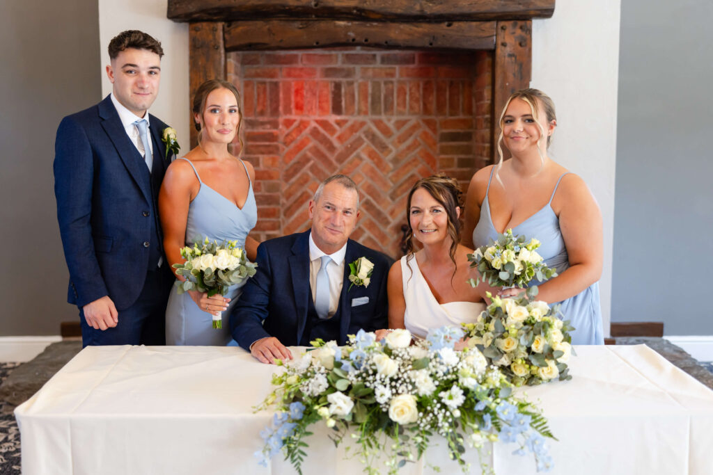 A bride and groom sit at a table with floral arrangements, flanked by two bridesmaids in light blue dresses and a man in a navy suit, posing for a wedding photo indoors. Copyright Somerside Photography Ltd // www.somersidephotography.com