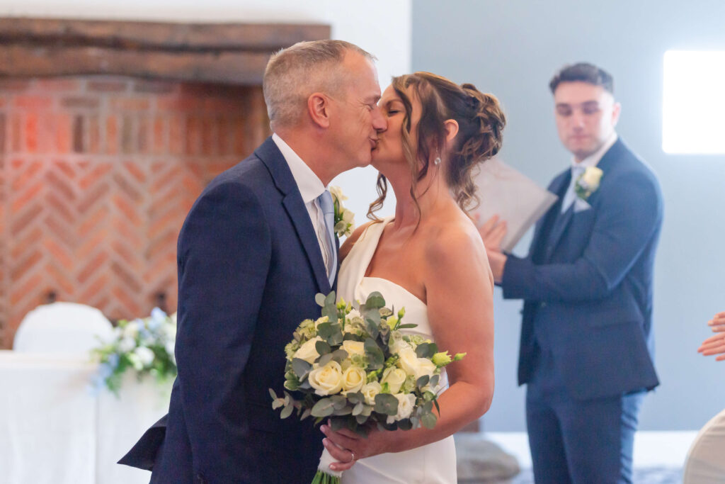 A bride and groom kiss at their wedding ceremony, holding a bouquet of flowers, whilst a man in a suit stands in the background holding papers. Copyright Somerside Photography Ltd // www.somersidephotography.com