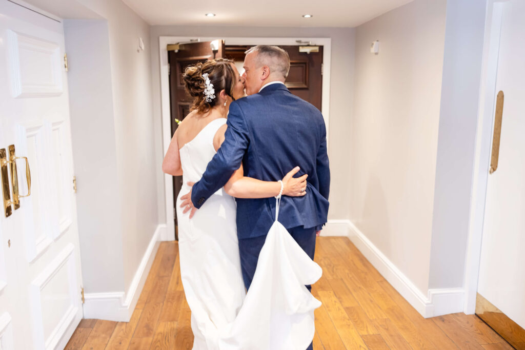 A bride and groom walk arm in arm down a corridor, with the groom holding the bride's dress train. They are dressed in wedding attire. Copyright Somerside Photography Ltd // www.somersidephotography.com
