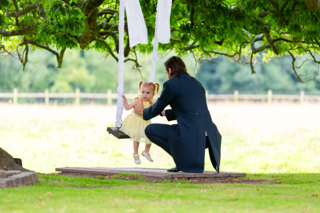 An adult in formal attire kneels beside a young child on a swing under a large tree in an outdoor setting. Copyright Somerside Photography Ltd // www.somersidephotography.com