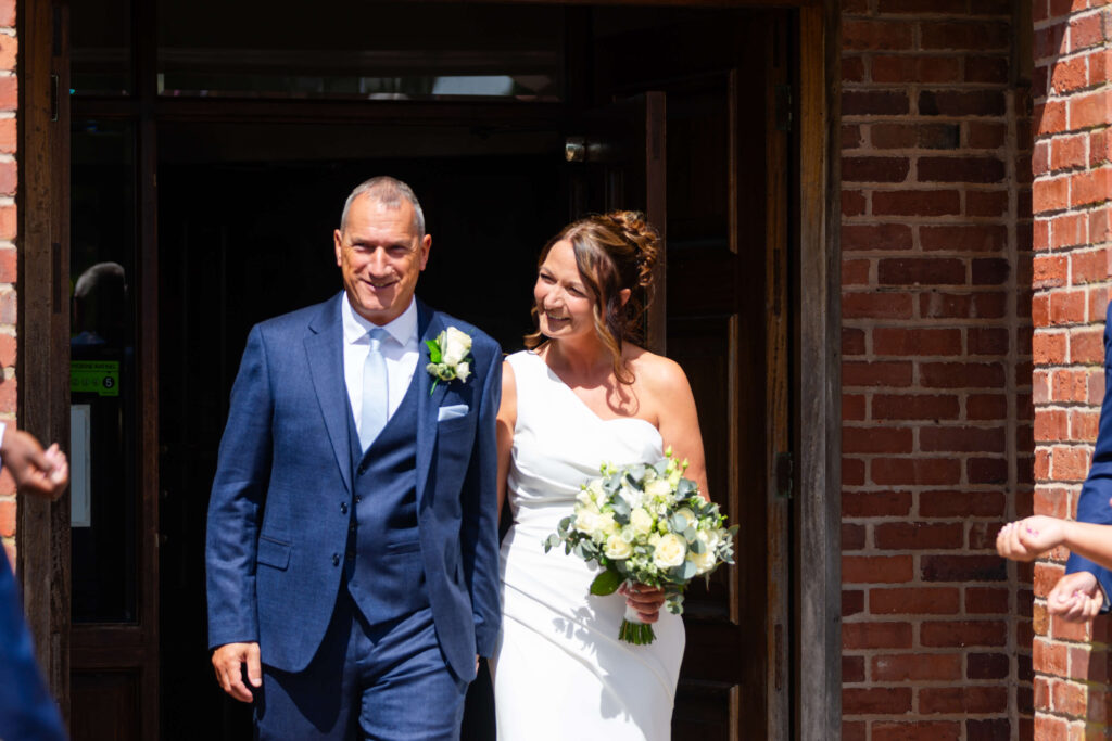A man in a blue suit and a woman in a white wedding dress holding a bunch of flowers walk out of a brick building, smiling in sunlight. Copyright Somerside Photography Ltd // www.somersidephotography.com