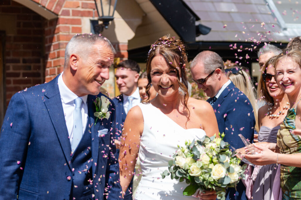A bride and groom walk outside surrounded by guests throwing confetti; the bride holds a bouquet and both are smiling. Copyright Somerside Photography Ltd // www.somersidephotography.com