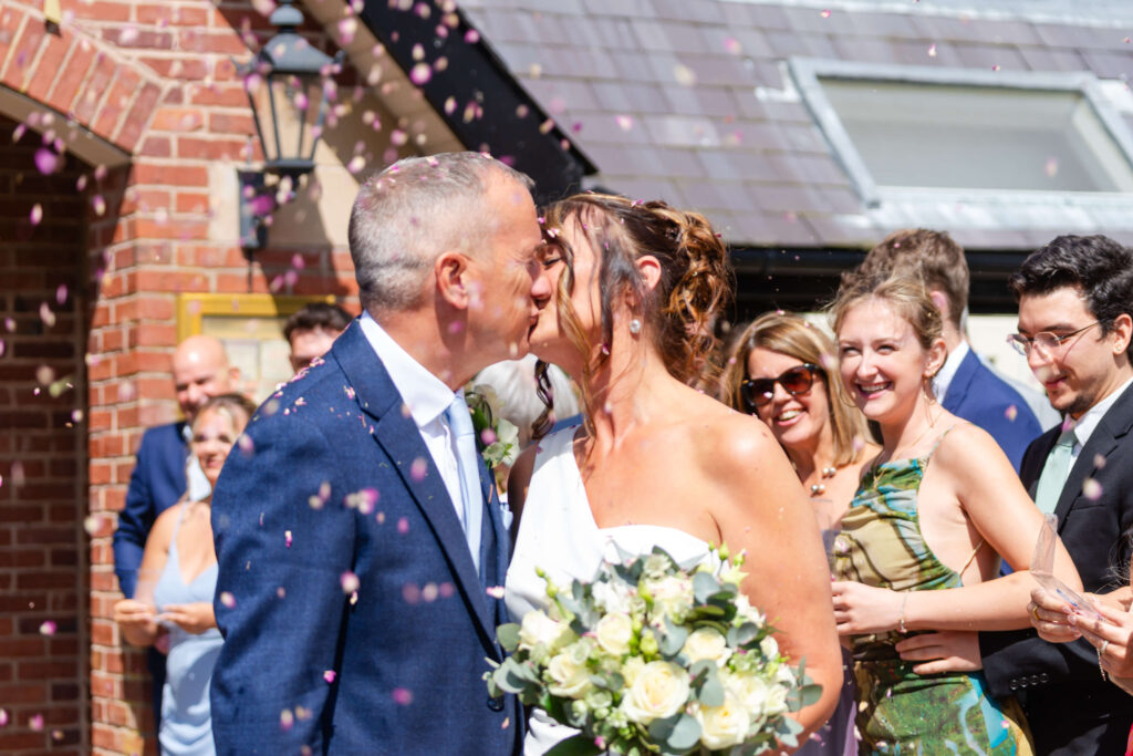 A bride and groom kiss outdoors whilst guests smile and throw flower petals. The bride holds a bouquet of white roses. Copyright Somerside Photography Ltd // www.somersidephotography.com