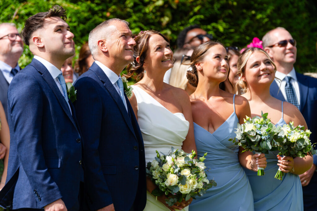 A wedding party poses outdoors, with the bride in a white dress and others in formal attire holding bouquets, all smiling and looking upwards. Copyright Somerside Photography Ltd // www.somersidephotography.com
