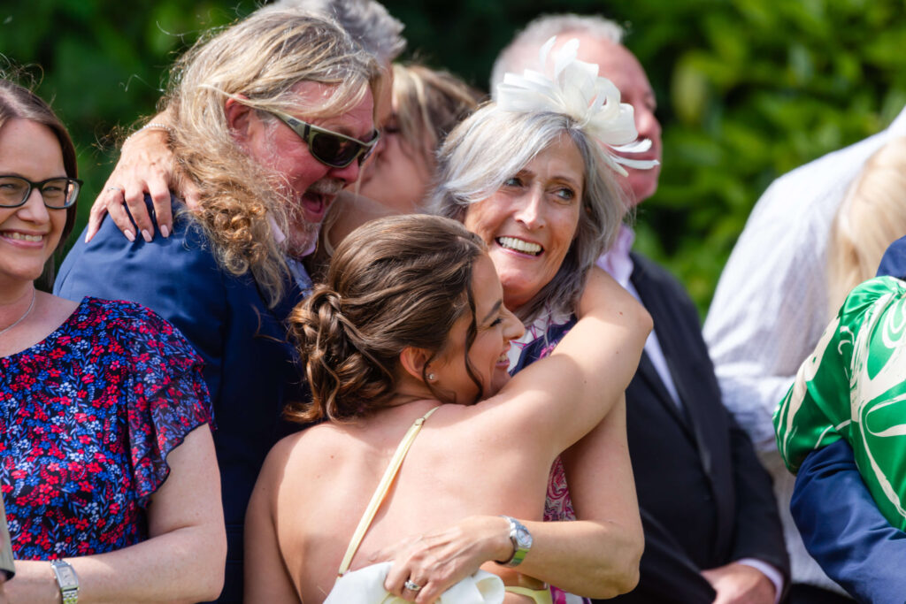 A woman in a white dress hugs an older woman in a light floral outfit and white fascinator, surrounded by smiling people outdoors. Copyright Somerside Photography Ltd // www.somersidephotography.com