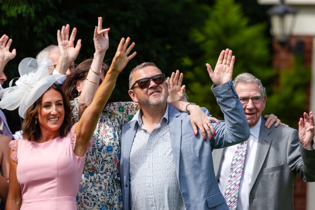 A group of people outdoors, smiling and raising their hands. One woman wears a white hat and pink dress; a man in sunglasses stands in the centre. Green foliage is in the background. Copyright Somerside Photography Ltd // www.somersidephotography.com