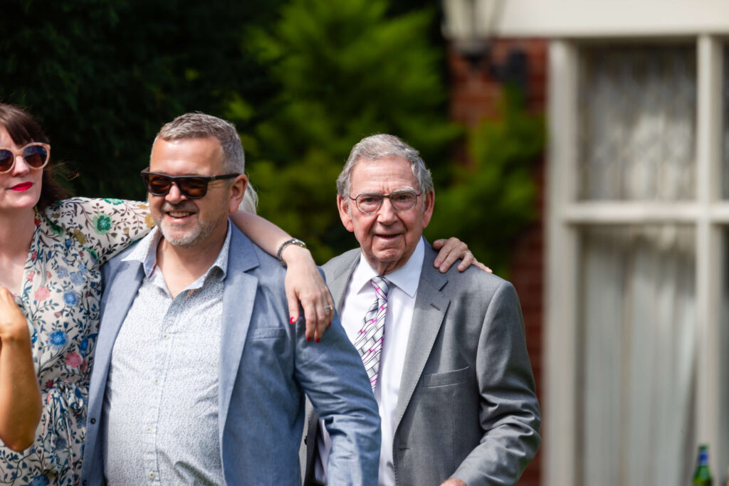 Three adults outdoors, two men in suits and sunglasses, with a woman partially visible in a floral dress, standing close together and smiling. Copyright Somerside Photography Ltd // www.somersidephotography.com