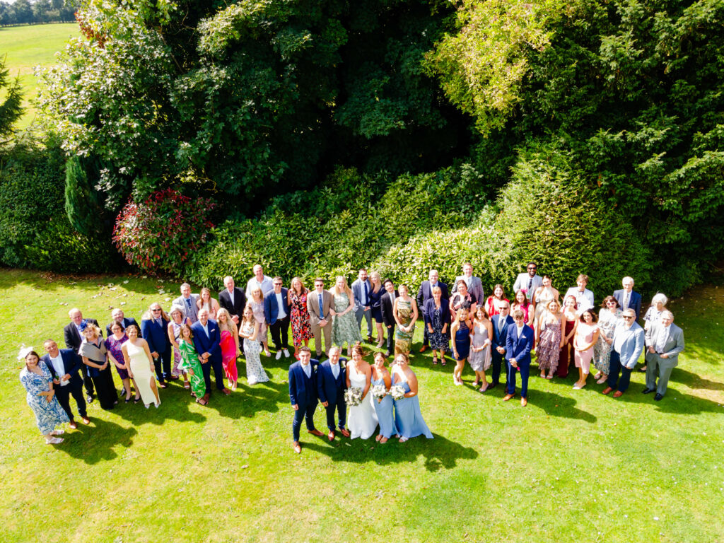 A group of people dressed in formal attire pose together on a grassy area near trees, likely for a wedding or formal event, viewed from above. Copyright Somerside Photography Ltd // www.somersidephotography.com