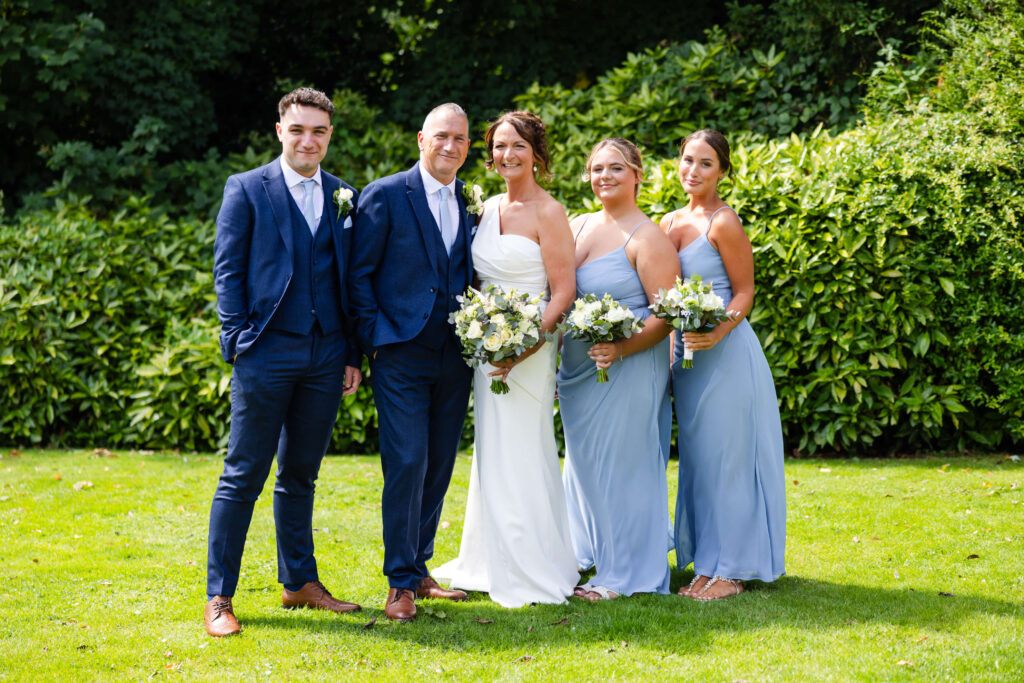 Five people dressed in formal attire stand on grass in front of green bushes, with three women holding bouquets and two men in suits, posing for a group photo. Copyright Somerside Photography Ltd // www.somersidephotography.com