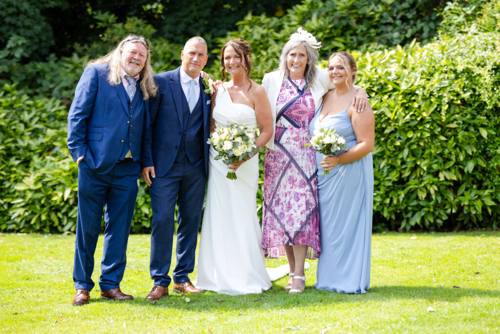 Five people are posing outdoors; two are wearing suits, two women are in dresses holding bouquets, and one woman is in a patterned dress and hat. They are standing on grass with greenery behind them. Copyright Somerside Photography Ltd // www.somersidephotography.com