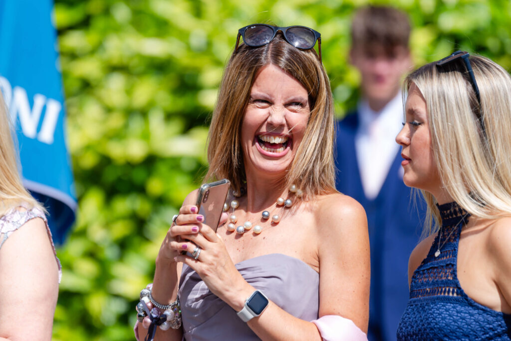 Woman in a strapless dress smiles broadly whilst holding a mobile, standing outdoors with others in sunny weather. Copyright Somerside Photography Ltd // www.somersidephotography.com