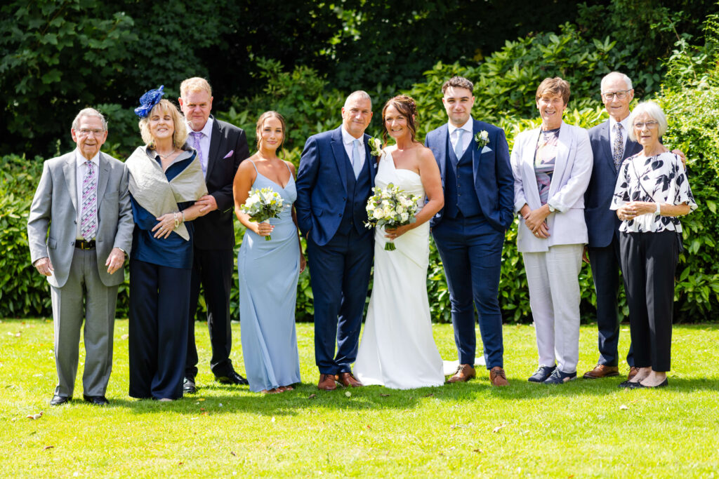 A wedding party poses outdoors on grass, with the bride and groom in the centre, flanked by bridesmaids, groomsmen, and older family members. Everyone is dressed formally. Copyright Somerside Photography Ltd // www.somersidephotography.com