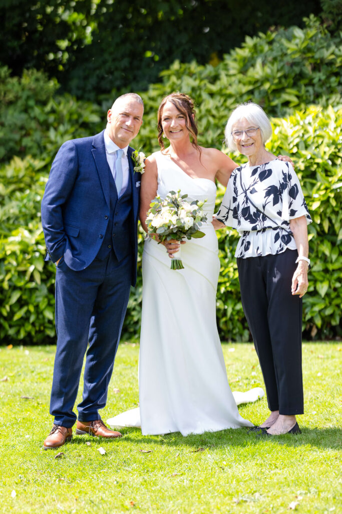 Three people pose outdoors: a man in a blue suit, a woman in a white one-shoulder wedding dress holding a bouquet, and an older woman in a black and white outfit. Copyright Somerside Photography Ltd // www.somersidephotography.com