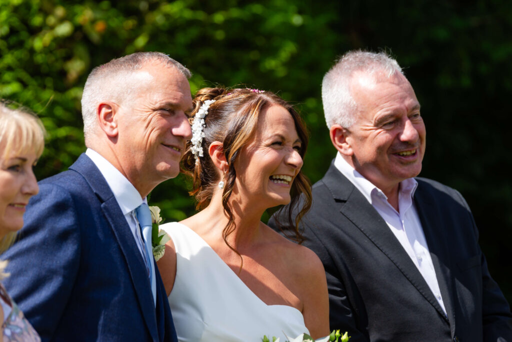 Three adults in formal attire smile outdoors on a sunny day. The woman in the centre wears a white dress and holds a bouquet, suggesting a wedding celebration. Trees are in the background. Copyright Somerside Photography Ltd // www.somersidephotography.com