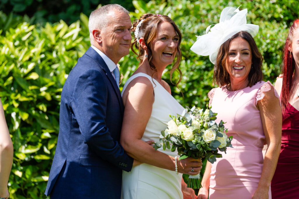 Three adults stand outdoors in bright sunlight; the woman in the centre wears a white wedding dress and holds a bouquet, while the others are dressed in formal attire and smiling. Copyright Somerside Photography Ltd // www.somersidephotography.com