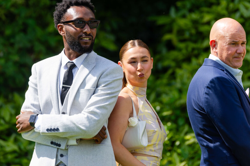 Three people stand outdoors; a man in a light suit with sunglasses, a woman in a patterned dress pulling a playful face, and a bald man in a dark suit, all facing slightly different directions. Copyright Somerside Photography Ltd // www.somersidephotography.com