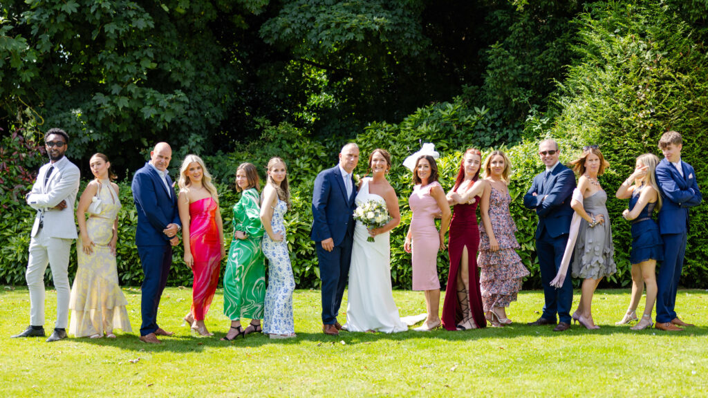 A wedding party stands in a row outdoors on green grass, with the bride and groom in the centre, surrounded by bridesmaids and groomsmen dressed in colourful attire. Copyright Somerside Photography Ltd // www.somersidephotography.com