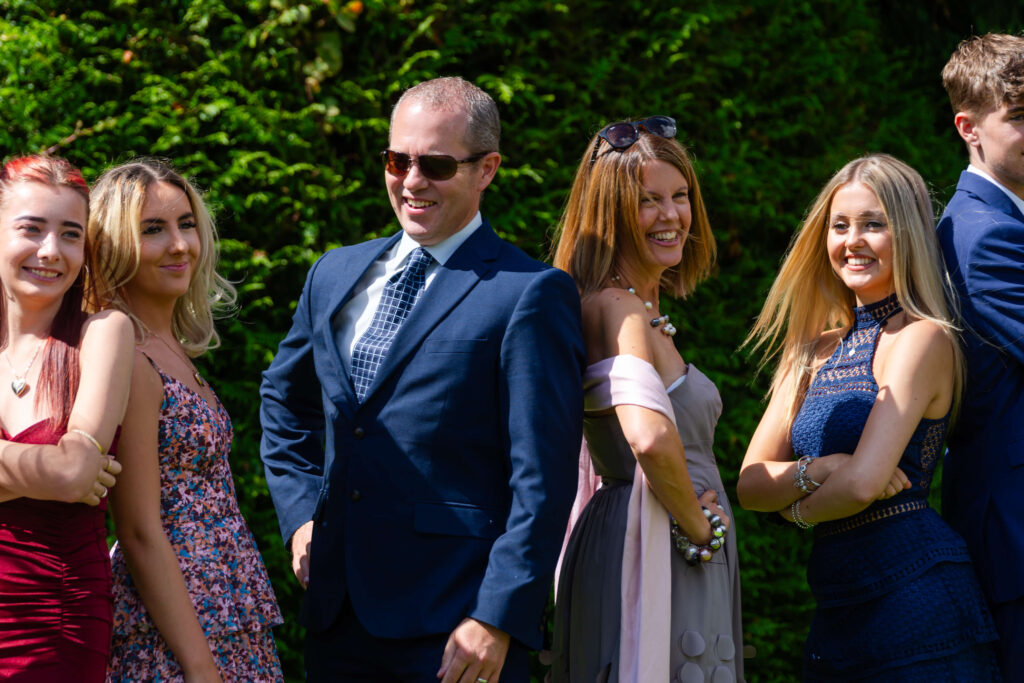A group of people dressed in formal attire stand outdoors with arms folded, smiling in the sunlight, with greenery in the background. Copyright Somerside Photography Ltd // www.somersidephotography.com