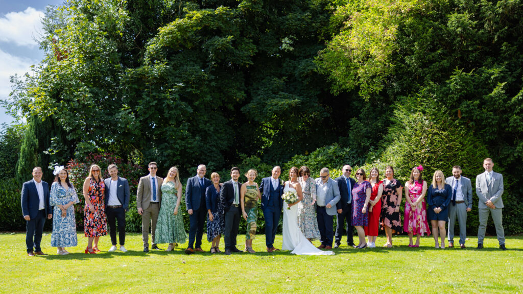 A group of people dressed in formal attire stands in a row on grass outdoors, with trees and greenery in the background. Copyright Somerside Photography Ltd // www.somersidephotography.com