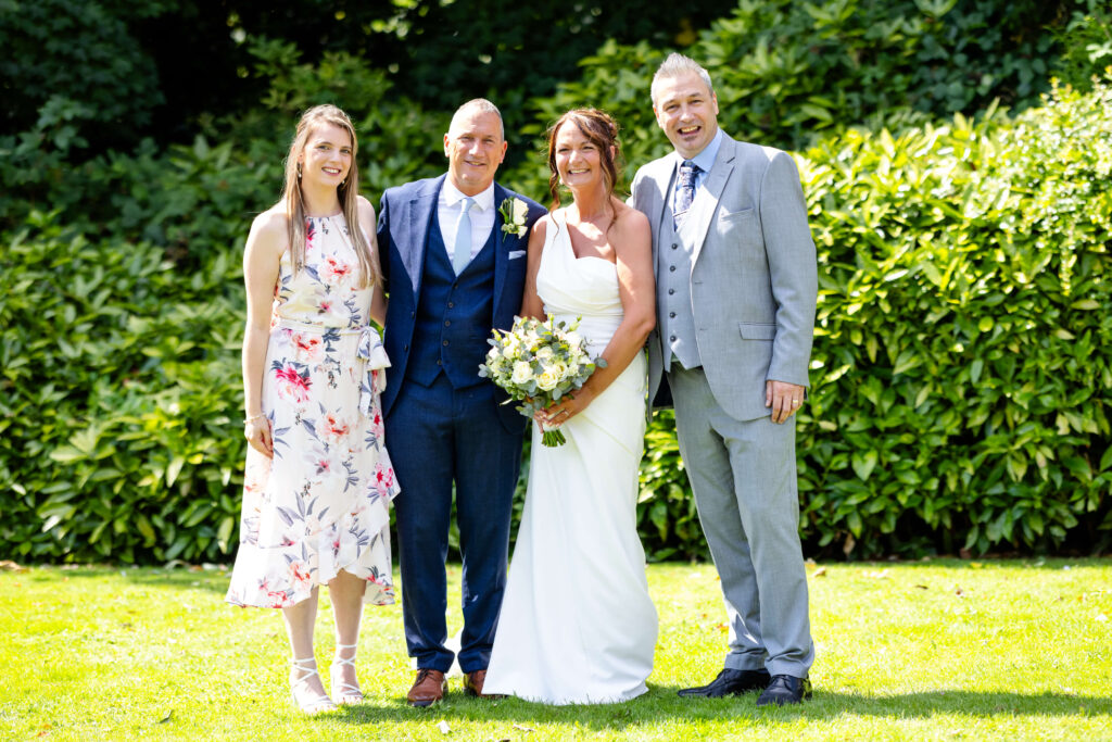 Four people pose for a photo outdoors, including a bride in a white dress holding a bouquet, a groom in a navy suit, and two other guests standing on grass in front of greenery. Copyright Somerside Photography Ltd // www.somersidephotography.com