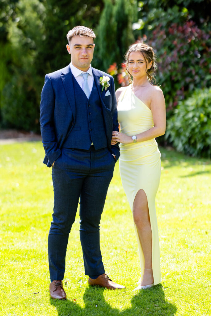 A man in a navy suit and a woman in a light yellow dress with a thigh-high slit pose together outdoors on a sunny day. Copyright Somerside Photography Ltd // www.somersidephotography.com