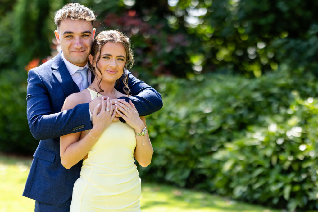 A man in a blue suit stands behind a woman in a yellow dress, embracing her from behind in a garden setting. Both are looking at the camera and smiling. Copyright Somerside Photography Ltd // www.somersidephotography.com