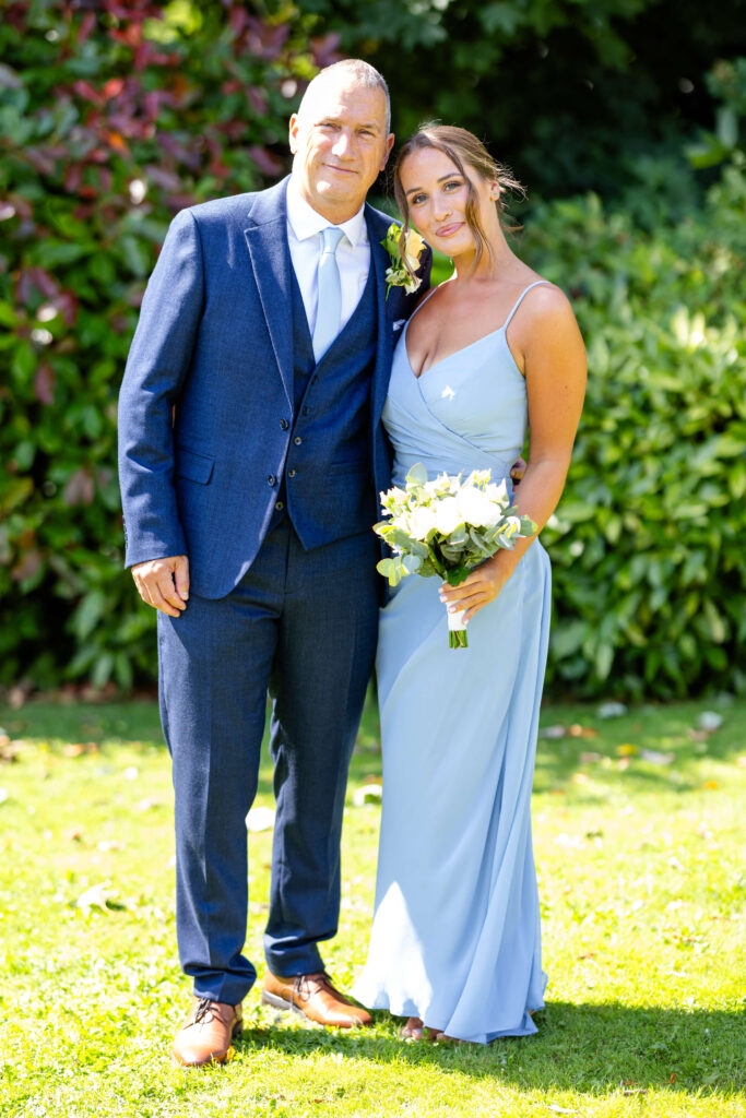 A man in a blue suit and a woman in a light blue dress holding a bouquet stand together on grass in front of greenery, posing for a photo. Copyright Somerside Photography Ltd // www.somersidephotography.com
