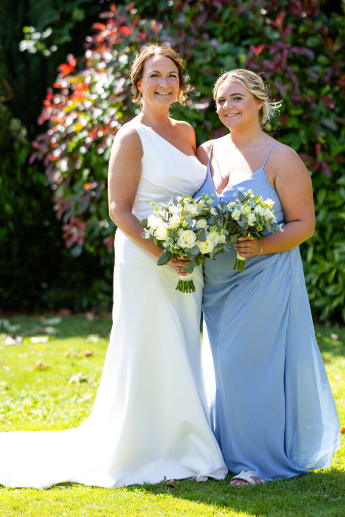 Two women in formal dresses, one in white and one in light blue, stand side by side holding flower bouquets outdoors on a sunny day. Copyright Somerside Photography Ltd // www.somersidephotography.com