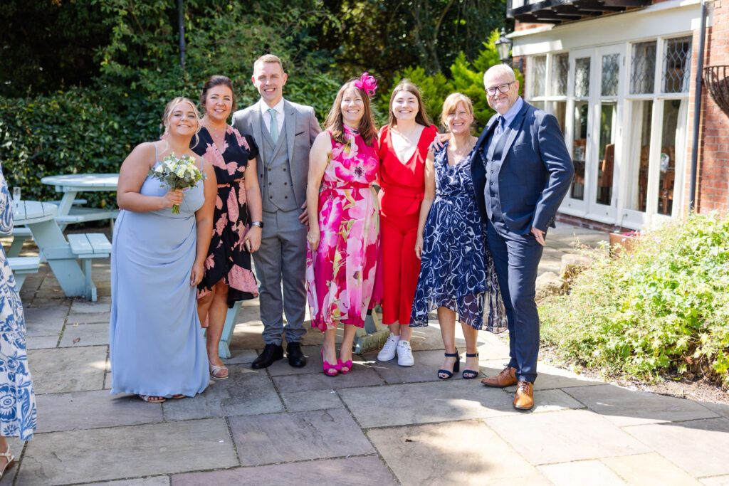 A group of seven adults dressed in formal and semi-formal attire pose together outdoors on a stone patio, smiling at the camera. Copyright Somerside Photography Ltd // www.somersidephotography.com