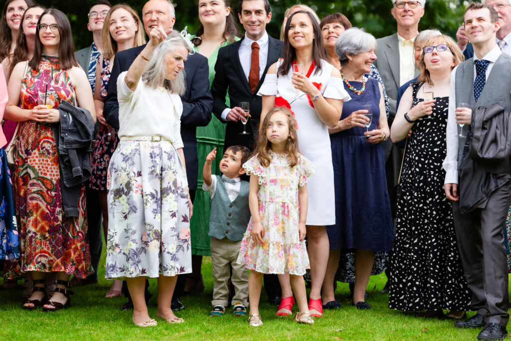 A group of people, including adults and children in formal attire, stand on grass outdoors, looking up and smiling. Copyright Somerside Photography Ltd // www.somersidephotography.com