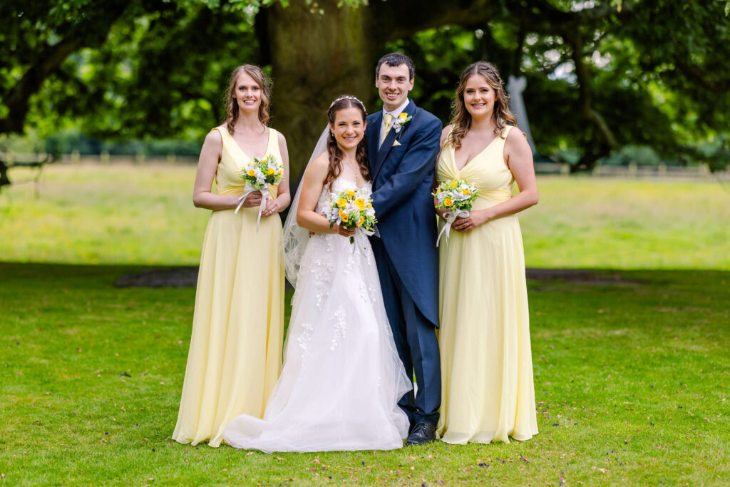 A bride and groom stand outdoors with two bridesmaids in yellow dresses, all holding bouquets, in front of a large tree. Copyright Somerside Photography Ltd // www.somersidephotography.com