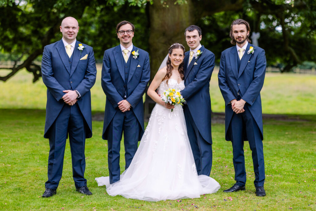 A bride in a white dress stands holding a bouquet, surrounded by four men in matching blue suits, posing outdoors on grass with trees in the background. Copyright Somerside Photography Ltd // www.somersidephotography.com