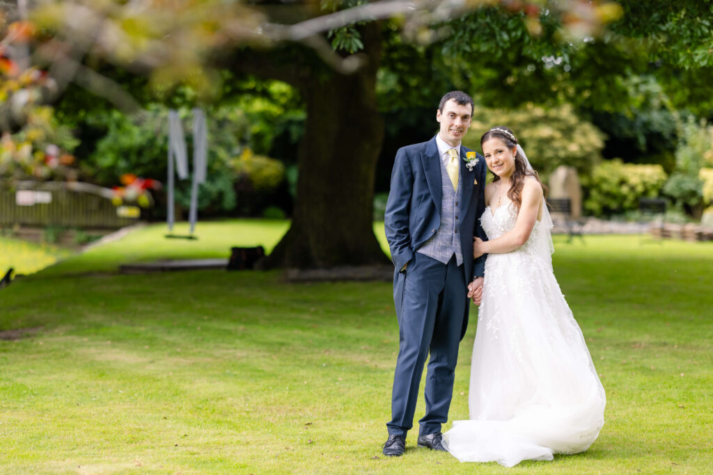 A bride and groom stand together on a grassy lawn in front of a large tree, posing for a wedding photo outdoors. Copyright Somerside Photography Ltd // www.somersidephotography.com
