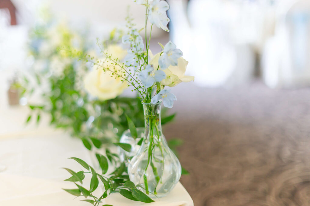 A small glass vase with light blue and white flowers and green foliage sits on a white table. Copyright Somerside Photography Ltd // www.somersidephotography.com