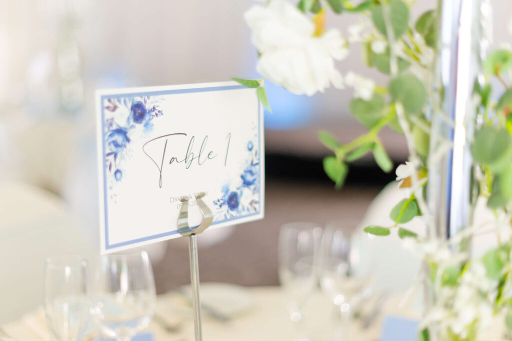 A table card labelled "Table 1" with blue floral decorations is displayed among green leaves and white flowers on a set dining table. Copyright Somerside Photography Ltd // www.somersidephotography.com