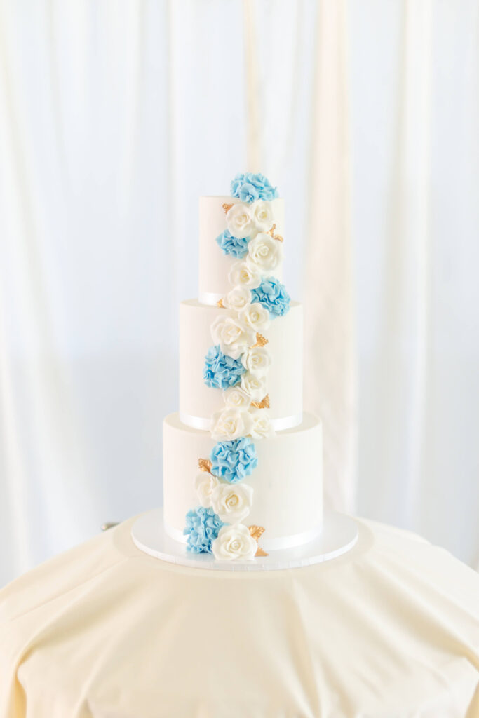 Three-tier white cake decorated with cascading blue and white flowers and small gold accents, displayed on a round white table against a light backdrop. Copyright Somerside Photography Ltd // www.somersidephotography.com