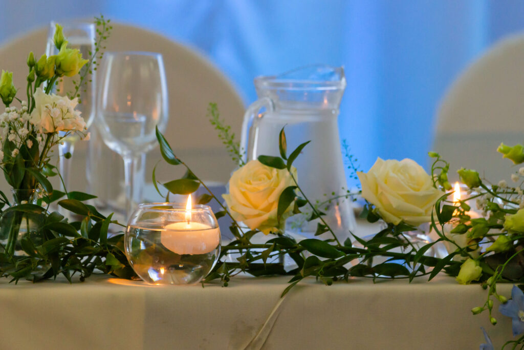 A wedding table centrepiece featuring white roses, greenery, floating candles in glass holders, a glass jug of water, and empty wine glasses on a white tablecloth. Copyright Somerside Photography Ltd // www.somersidephotography.com
