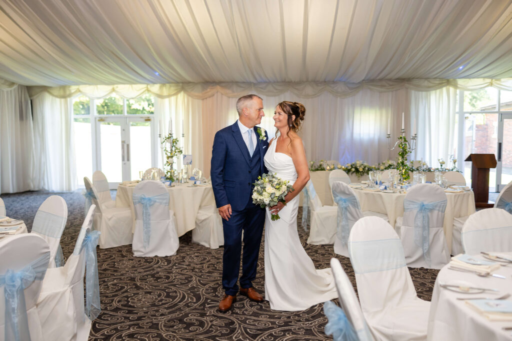 A bride and groom stand together smiling in a decorated wedding reception venue with round tables and white chairs. Copyright Somerside Photography Ltd // www.somersidephotography.com